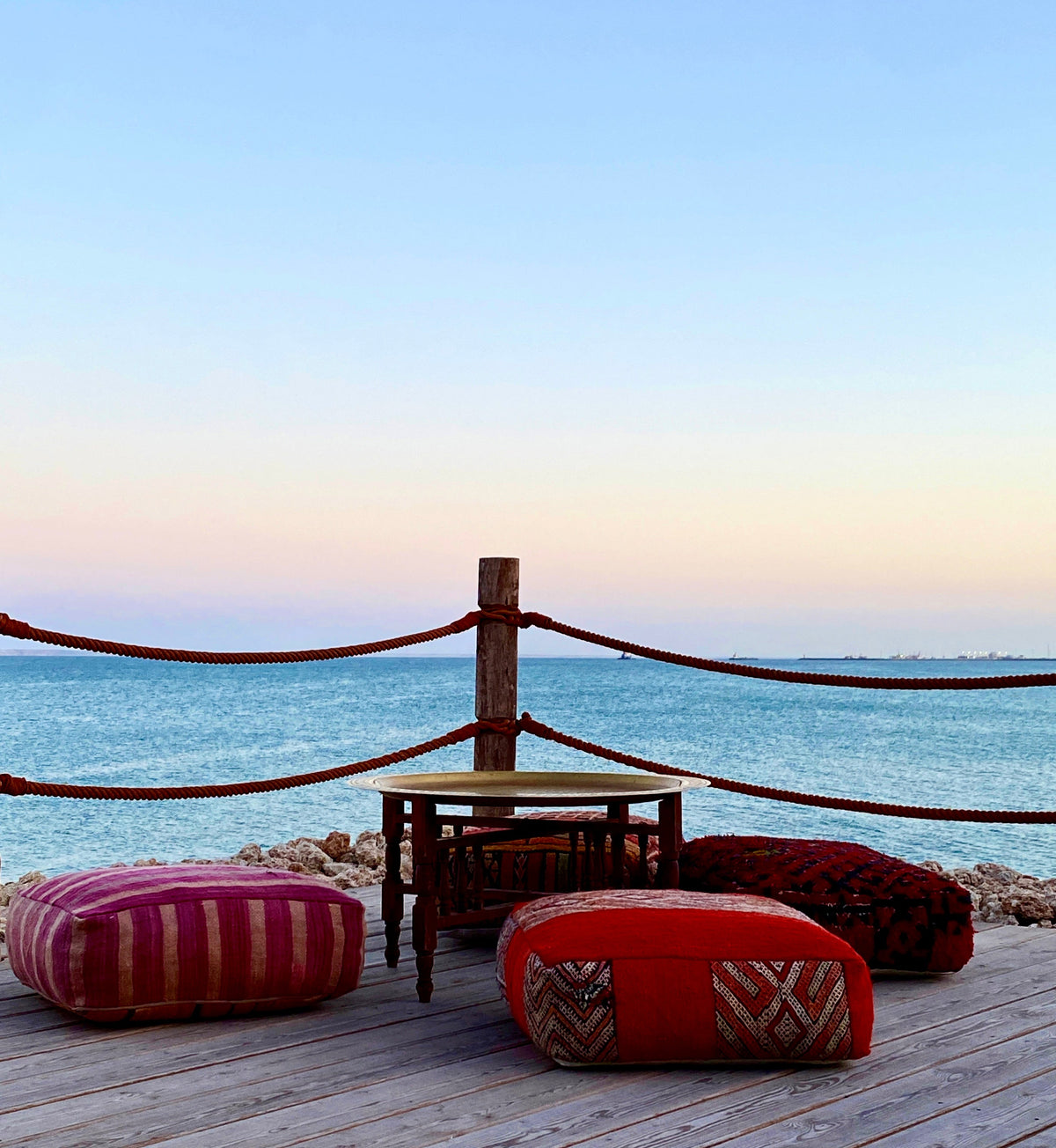 a trip of artisan hand-crafted moroccan cushions around an artisan hand-crafted tea tray on a deck over looking the lagoon in Dakhla during sunset