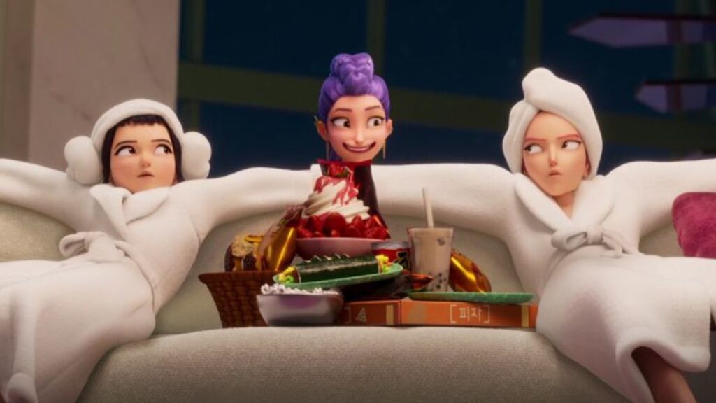 Netflix's Kpop Demon Hunters movie image of the three main characters Zoe and Mira sitting on the sofa relaxing in white robes and slippers with their hair wrapped in a towel while Rumi with her make up on and purple hair braided is sneaking between the two to tell them they are going back to work.