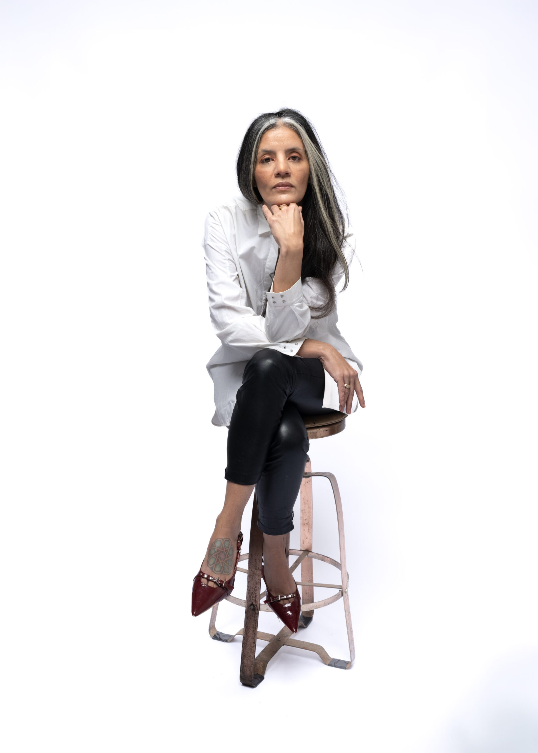 Woman with a streak of grey in her hair leaning into the camera with her hand on her chin and elbow resting on her lap in a white button down men's dress shirt sitting on a stool with her legs crossed and red kitten heels on.