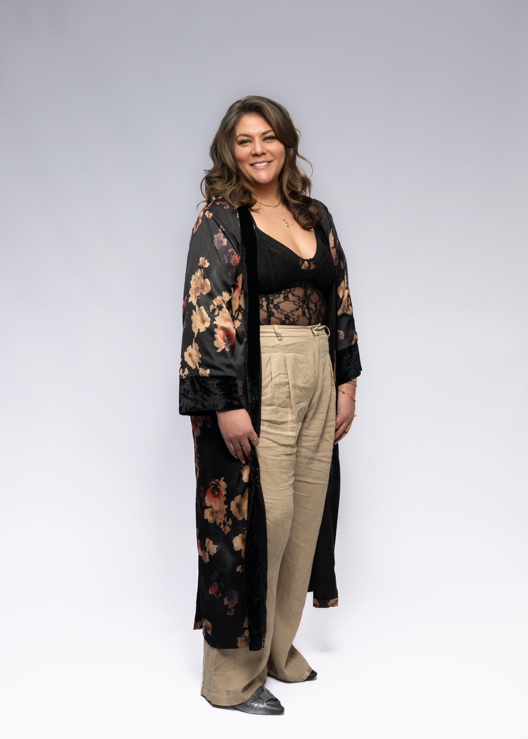 A a woman with long curled brown hair and tan skin is smiling directly into the camera wearing a black Kim + Ono kimono and black tank top and beige linen pants standing and smiling at the camera